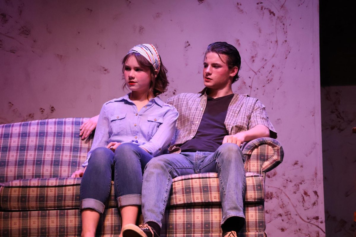 Sandy (Isabella Walter) and Sodapop ( Grant Timmons) Sit together on the couch in the beginning scenes of The Outsiders.