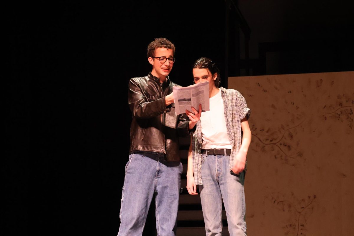 Twobit (Andrew Busack) shows Ponyboy (Sam Hannig) the newspaper to see that he is seen as a hero for saving the kids in the church.